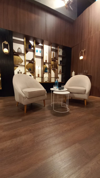 Dos sillones modernos de tapizado beige claro con patas de madera, dispuestos en un área de espera con suelo de madera oscura. Entre ellos hay dos mesas pequeñas redondas, una con una botella y vaso de agua. Al fondo se observa una pared decorativa con estanterías iluminadas y un gran logo dorado de MasterChef