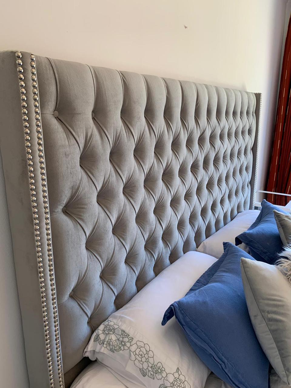 Respaldo de cama tapizado en tela gris con diseño capitoné y tachas decorativas en los bordes, acompañado de almohadones decorativos y ropa de cama en tonos azules