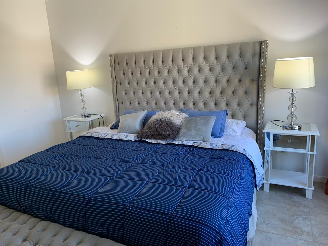 Respaldo de cama tapizado en tela gris con diseño capitoné y tachas decorativas en los bordes, acompañado de almohadones decorativos y ropa de cama en tonos azules