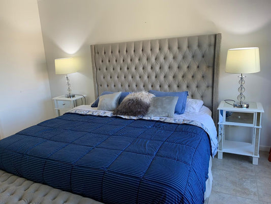Respaldo de cama tapizado en tela gris con diseño capitoné y tachas decorativas en los bordes, acompañado de almohadones decorativos y ropa de cama en tonos azules
