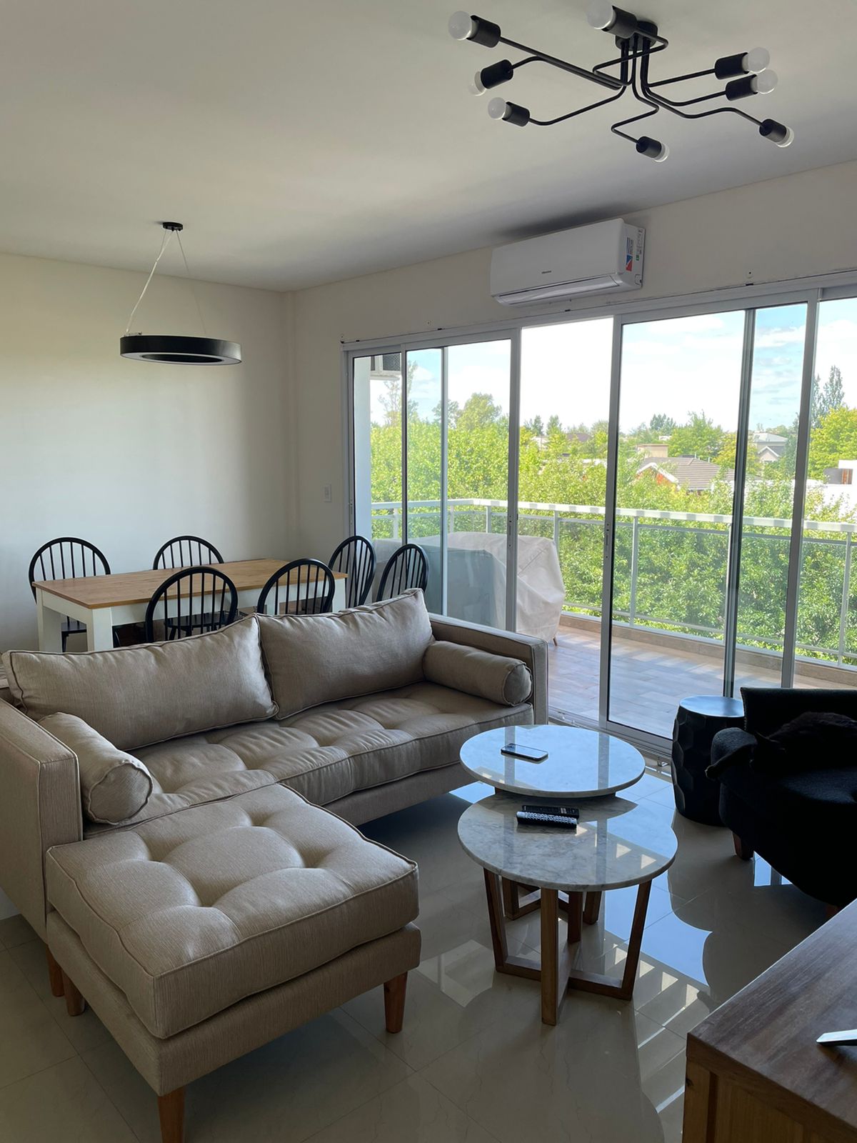 Sofá esquinero tapizado en tono beige claro con chaise longue y patas de madera, de diseño moderno y confortable, ideal para un living luminoso con amplios ventanales