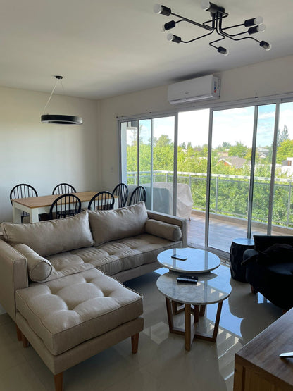 Sofá esquinero tapizado en tono beige claro con chaise longue y patas de madera, de diseño moderno y confortable, ideal para un living luminoso con amplios ventanales