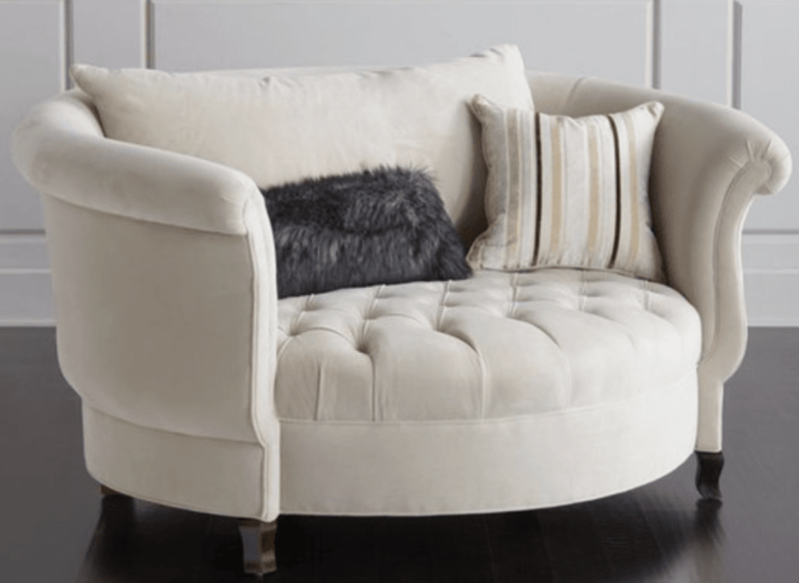 Sillón redondo de estilo elegante en color crema, con respaldo curvo envolvente, apoyabrazos enrollados y asiento capitoné, patas cortas de madera.