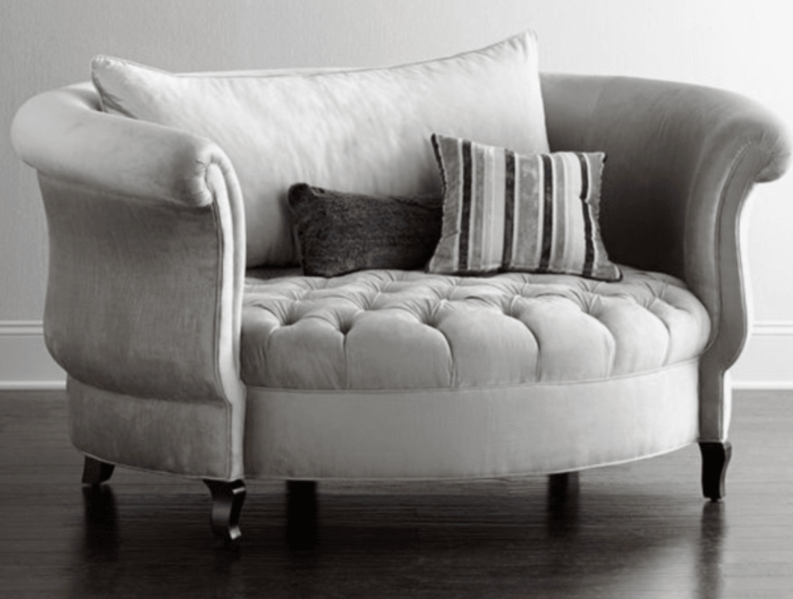 Sillón redondo de estilo elegante en color gris claro, con respaldo curvo envolvente, apoyabrazos enrollados y asiento capitoné, patas cortas de madera.