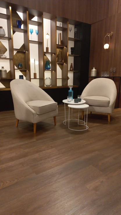 Dos sillones modernos de tapizado beige claro con patas de madera, dispuestos en un área de espera con suelo de madera oscura. Entre ellos hay dos mesas pequeñas redondas, una con una botella y vaso de agua. Al fondo se observa una pared decorativa con estanterías iluminadas y un gran logo dorado de MasterChef