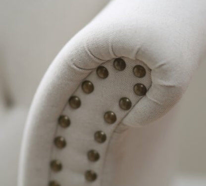 Sillón estilo clásico tapizado en color gris claro, con respaldo capitoné alto, orejas curvas, apoyabrazos redondeados y tachas decorativas, patas de madera talladas