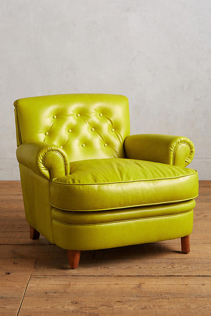 Sillón individual tapizado en color oliva, con respaldo acolchado decorado con botones capitoné, apoyabrazos redondeados y patas cortas de madera en tono natural.