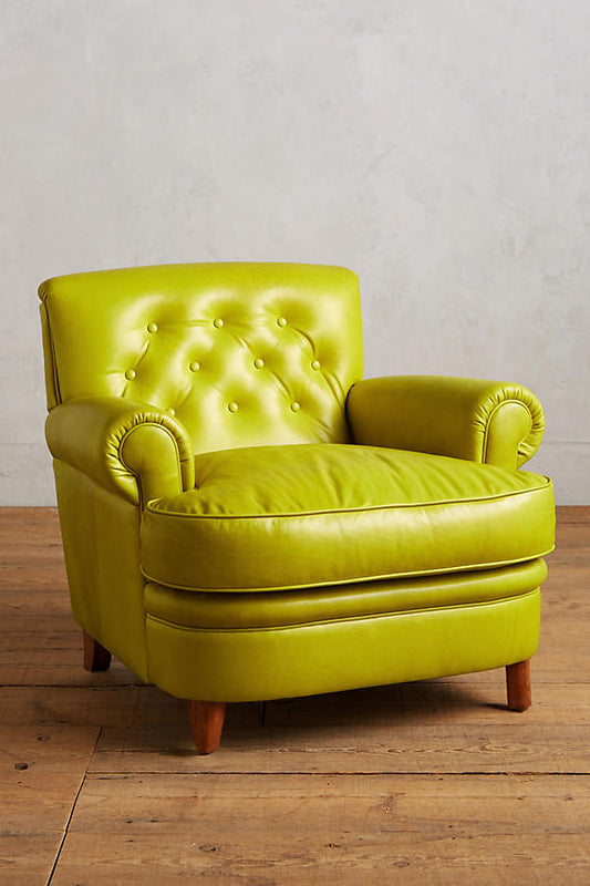Sillón individual tapizado en color oliva, con respaldo acolchado decorado con botones capitoné, apoyabrazos redondeados y patas cortas de madera en tono natural.
