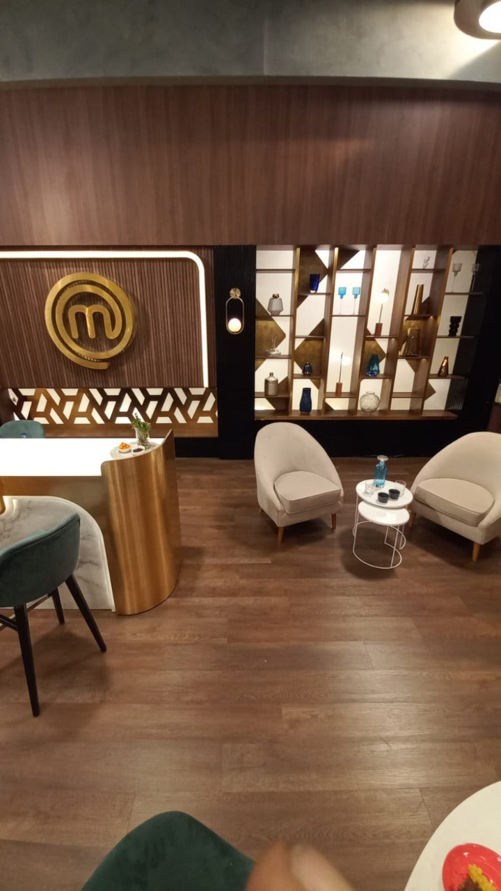Dos sillones modernos de tapizado beige claro con patas de madera, dispuestos en un área de espera con suelo de madera oscura. Entre ellos hay dos mesas pequeñas redondas, una con una botella y vaso de agua. Al fondo se observa una pared decorativa con estanterías iluminadas y un gran logo dorado de MasterChef