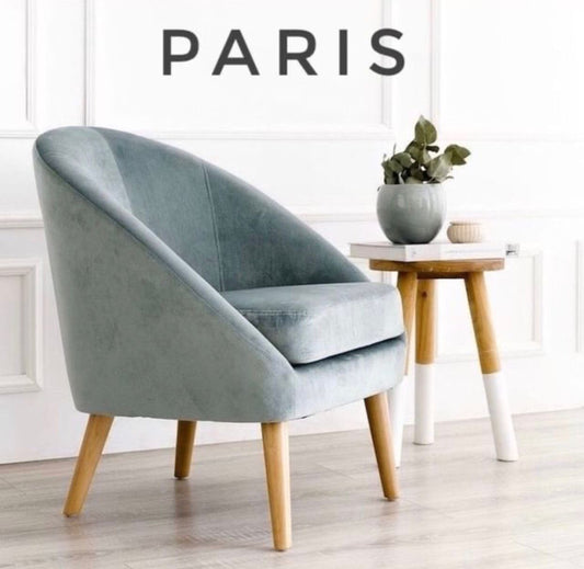 Sillón Paris de un cuerpo tapizado en celeste suave, con forma redondeada y apoyabrazos integrados, sobre piso de madera y alfombra texturada en living luminoso. Tienda de Banquitos.