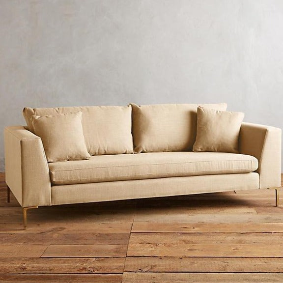 Sillón Bélgica de dos cuerpos con estructura recta, patas metálicas doradas finas, respaldo con almohadones sueltos y diseño atemporal. Estilo minimalista y acogedor.