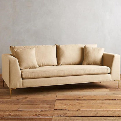 Sillón Bélgica de dos cuerpos con estructura recta, patas metálicas doradas finas, respaldo con almohadones sueltos y diseño atemporal. Estilo minimalista y acogedor.