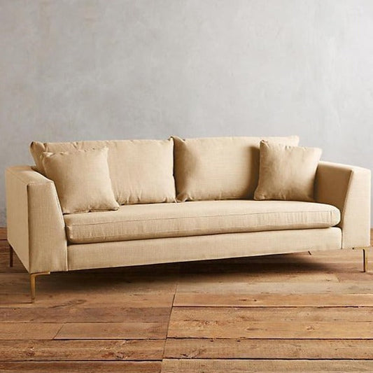 Sillón Bélgica de dos cuerpos con estructura recta, patas metálicas doradas finas, respaldo con almohadones sueltos y diseño atemporal. Estilo minimalista y acogedor.