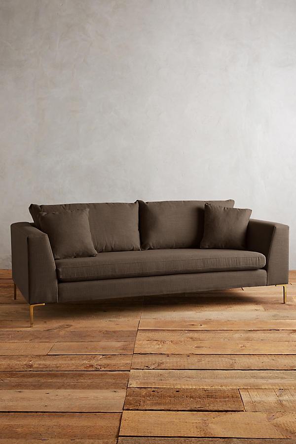 Sillón Bélgica de dos cuerpos con estructura recta, patas metálicas doradas finas, respaldo con almohadones sueltos y diseño atemporal. Estilo minimalista y acogedor.