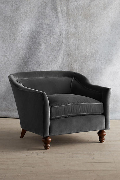 Sillón individual tapizado en color gris, con diseño de respaldo envolvente, asiento amplio y patas torneadas de madera oscura.