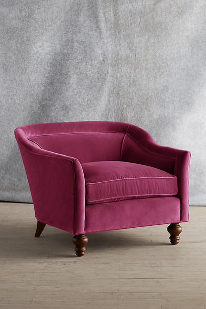 Sillón individual tapizado en color magenta, con diseño de respaldo envolvente, asiento amplio y patas torneadas de madera oscura.