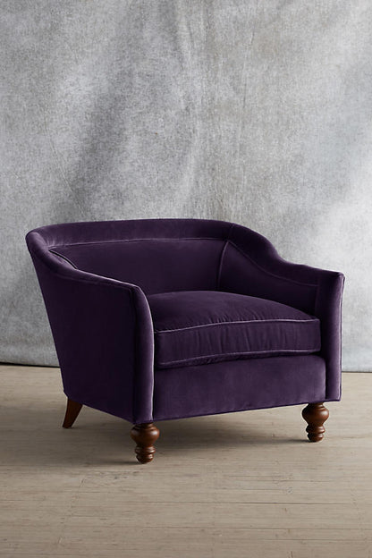 Sillón individual tapizado en color violeta, con diseño de respaldo envolvente, asiento amplio y patas torneadas de madera oscura.