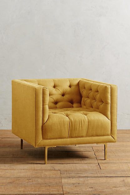 Sillón individual tapizado en amarillo, de forma cúbica con respaldo y brazos rectos, capitoné de botones en todo el interior y patas delgadas de madera.