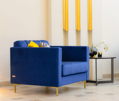 Sillón de un cuerpo tapizado en azul, con respaldo recto acanalado, apoyabrazos rectangulares y base baja con patas doradas.