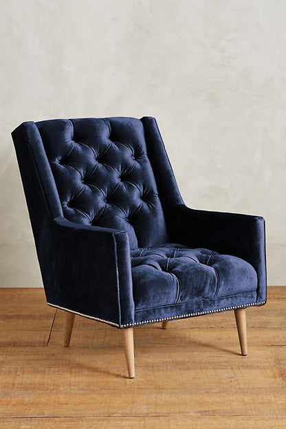 Sillón individual tapizado en azul, con respaldo capitoné de botones, brazos rectos y patas de madera inclinadas.