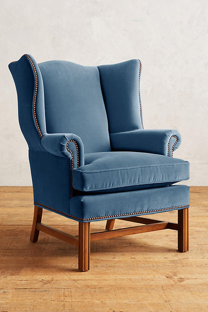 Sillón individual tapizado en terciopelo celeste, con respaldo alto y orejero, cojines sueltos en asiento y respaldo, y base de madera vista con patas cruzadas.