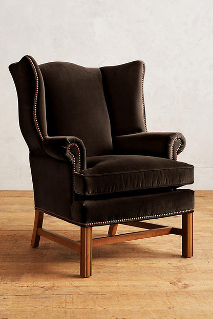 Sillón individual tapizado en terciopelo marron, con respaldo alto y orejero, cojines sueltos en asiento y respaldo, y base de madera vista con patas cruzadas.