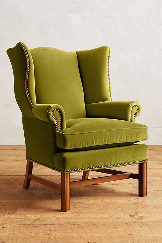 Sillón individual tapizado en terciopelo verde, con respaldo alto y orejero, cojines sueltos en asiento y respaldo, y base de madera vista con patas cruzadas.