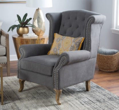 Sillón estilo clásico tapizado en color gris claro, con respaldo capitoné alto, orejas curvas, apoyabrazos redondeados y tachas decorativas, patas de madera talladas