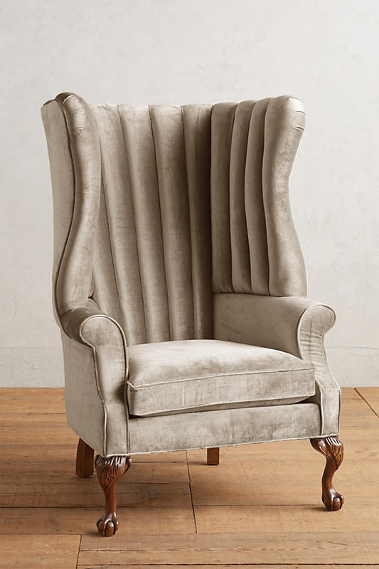 Sillón individual tapizado en beige, con respaldo alto acanalado de líneas verticales, brazos curvos y patas delanteras torneadas con ruedas pequeñas.