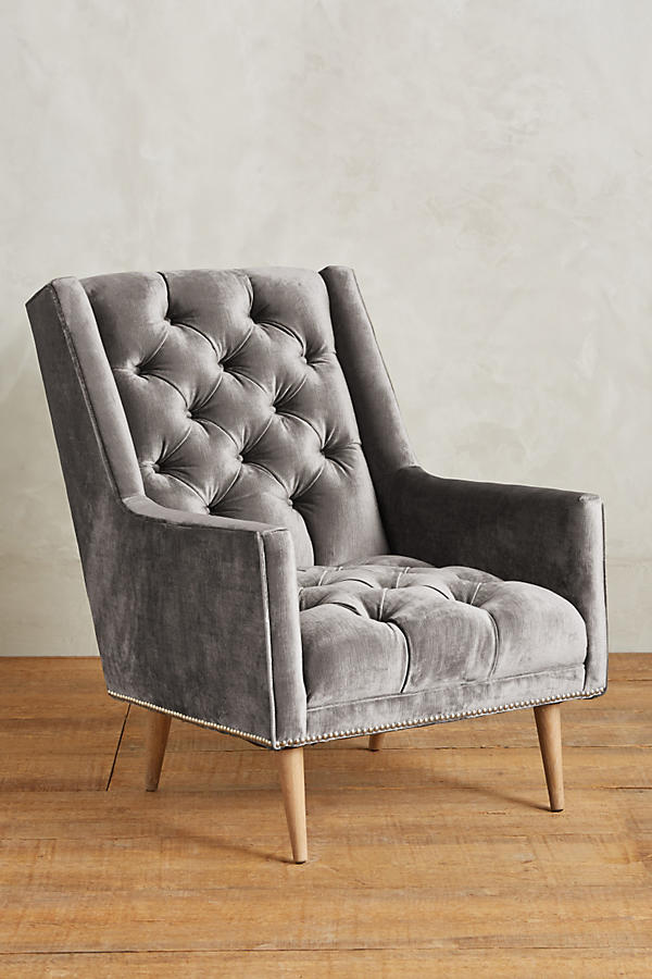 Sillón individual tapizado en gris, con respaldo capitoné de botones, brazos rectos y patas de madera inclinadas.