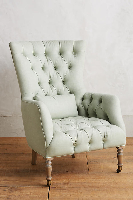 Sillón individual tapizado en gris, con respaldo alto y capitoné, cojín lumbar suelto, brazos curvos y patas delanteras torneadas en madera clara.