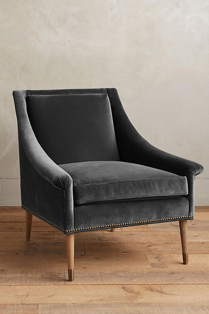 Sillón individual tapizado en gris claro, de líneas rectas con respaldo y asiento acolchados, brazos delgados y patas de madera rectas con detalle de tachas decorativas en la parte inferior.