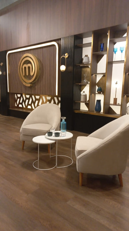 Dos sillones modernos de tapizado beige claro con patas de madera, dispuestos en un área de espera con suelo de madera oscura. Entre ellos hay dos mesas pequeñas redondas, una con una botella y vaso de agua. Al fondo se observa una pared decorativa con estanterías iluminadas y un gran logo dorado de MasterChef