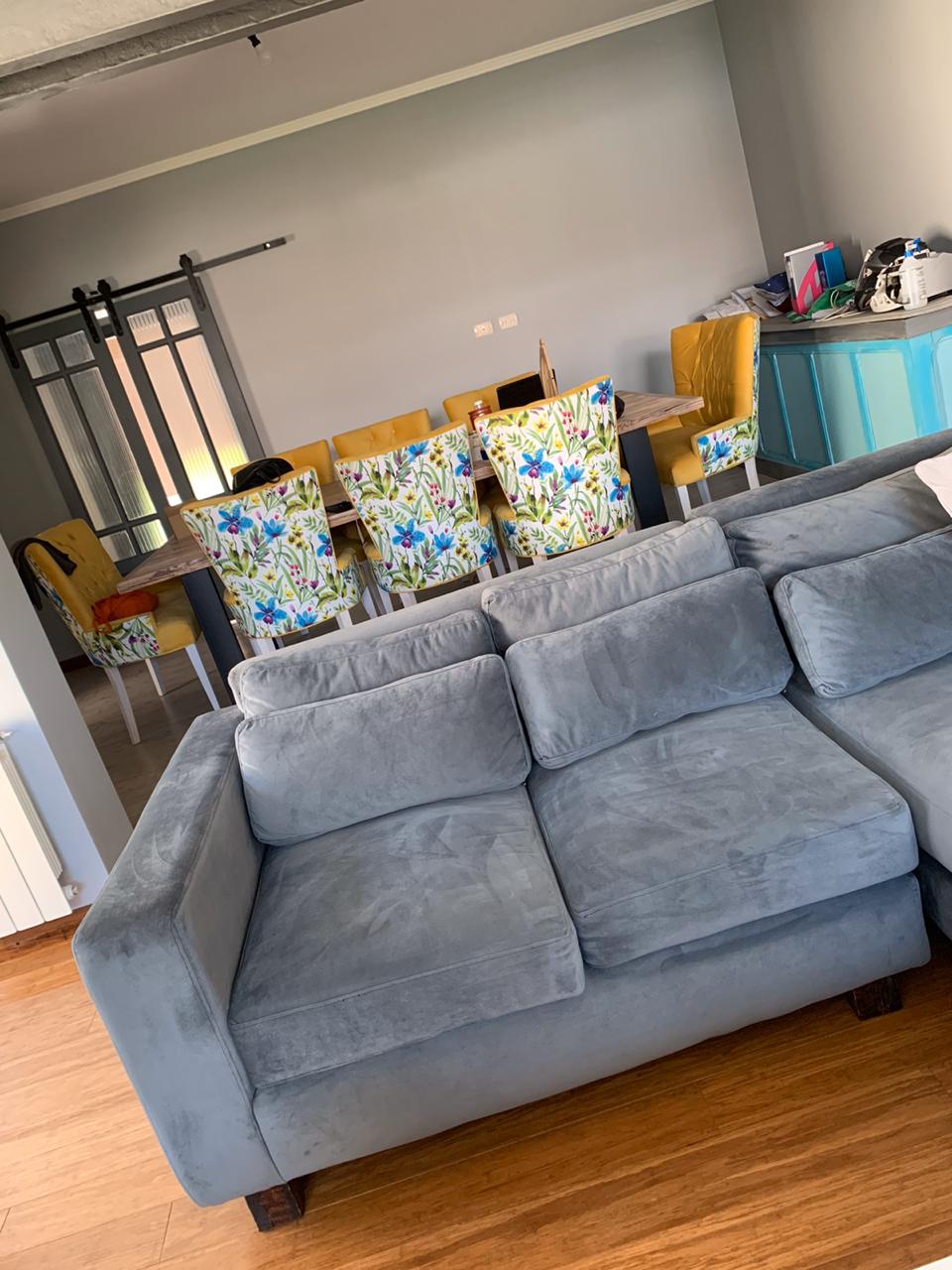 Sillón esquinero de tres cuerpos en tapizado gris, con cojines amplios y chaise longue, ubicado sobre piso de madera en un living moderno
