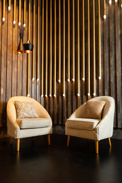 Par de sillones individuales tapizados en tono beige con patas de madera, de diseño moderno y respaldo curvo, decorados con almohadones a tono, ideales para un ambiente elegante y acogedor