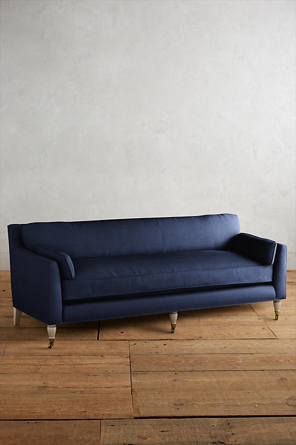 Fotografía de diseño de interiores estilo editorial que muestra un living moderno e industrial sofisticado. En el centro se destaca un sillón de tres cuerpos tapizado en lino gris claro con patas torneadas blancas y rueditas de bronce, idéntico al modelo de referencia. Está ubicado sobre una alfombra de diseño clásico envejecido superpuesta a una gran alfombra de yute