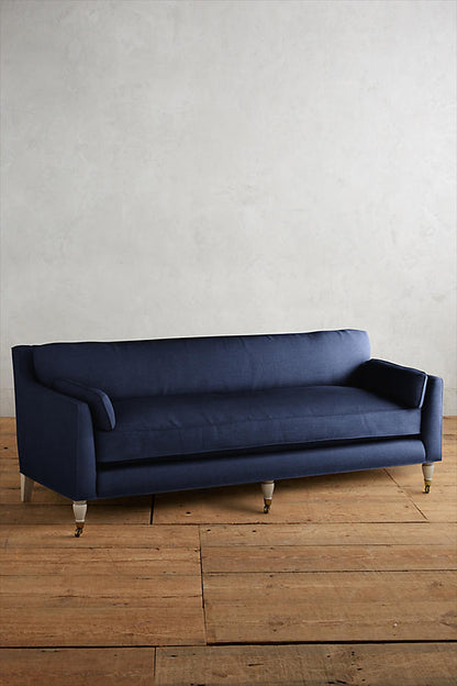 Fotografía de diseño de interiores estilo editorial que muestra un living moderno e industrial sofisticado. En el centro se destaca un sillón de tres cuerpos tapizado en lino gris claro con patas torneadas blancas y rueditas de bronce, idéntico al modelo de referencia. Está ubicado sobre una alfombra de diseño clásico envejecido superpuesta a una gran alfombra de yute