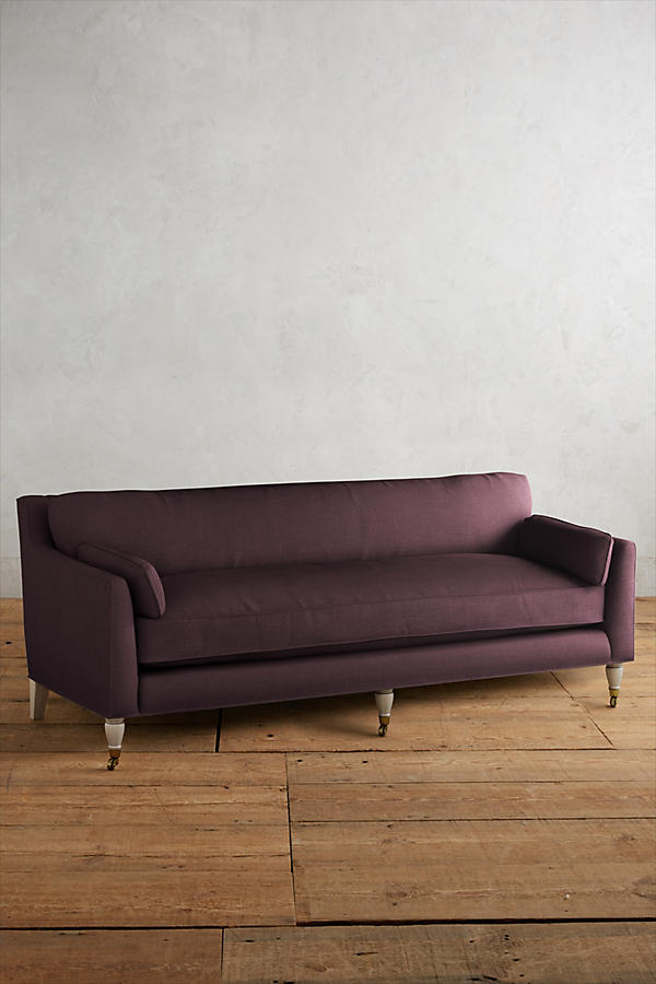 Fotografía de diseño de interiores estilo editorial que muestra un living moderno e industrial sofisticado. En el centro se destaca un sillón de tres cuerpos tapizado en lino gris claro con patas torneadas blancas y rueditas de bronce, idéntico al modelo de referencia. Está ubicado sobre una alfombra de diseño clásico envejecido superpuesta a una gran alfombra de yute