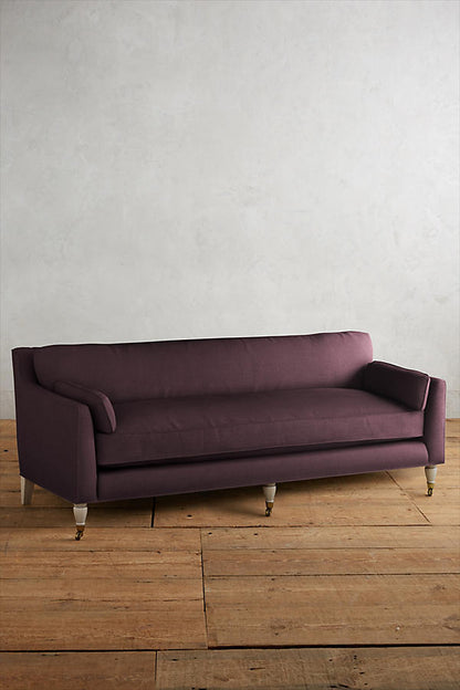 Fotografía de diseño de interiores estilo editorial que muestra un living moderno e industrial sofisticado. En el centro se destaca un sillón de tres cuerpos tapizado en lino gris claro con patas torneadas blancas y rueditas de bronce, idéntico al modelo de referencia. Está ubicado sobre una alfombra de diseño clásico envejecido superpuesta a una gran alfombra de yute