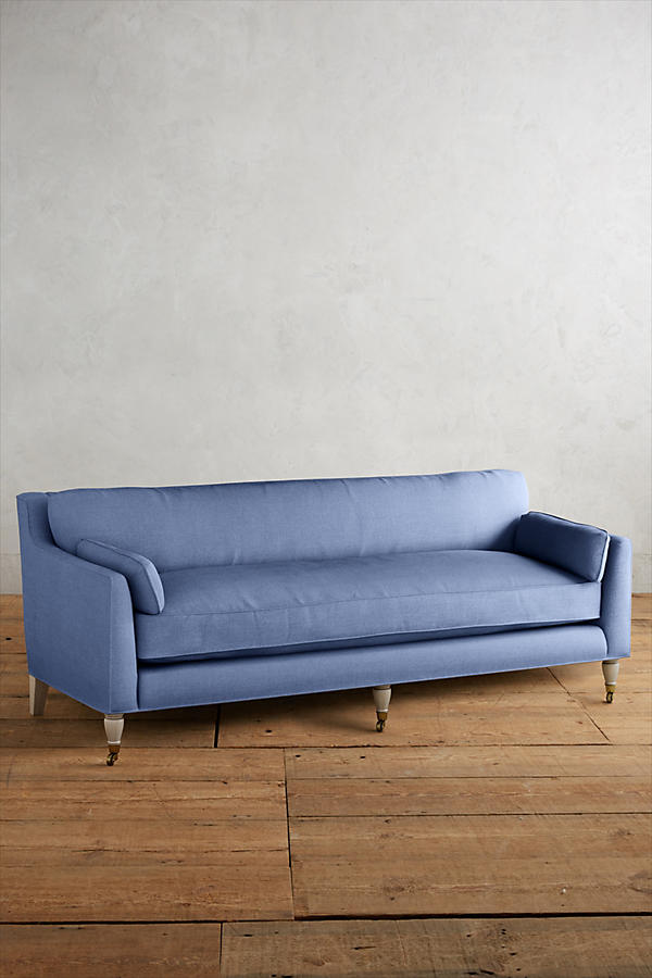 Fotografía de diseño de interiores estilo editorial que muestra un living moderno e industrial sofisticado. En el centro se destaca un sillón de tres cuerpos tapizado en lino gris claro con patas torneadas blancas y rueditas de bronce, idéntico al modelo de referencia. Está ubicado sobre una alfombra de diseño clásico envejecido superpuesta a una gran alfombra de yute