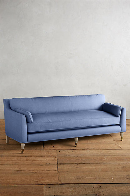 Fotografía de diseño de interiores estilo editorial que muestra un living moderno e industrial sofisticado. En el centro se destaca un sillón de tres cuerpos tapizado en lino gris claro con patas torneadas blancas y rueditas de bronce, idéntico al modelo de referencia. Está ubicado sobre una alfombra de diseño clásico envejecido superpuesta a una gran alfombra de yute