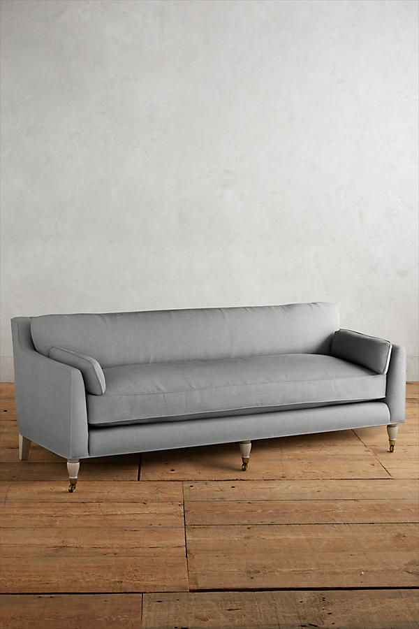 Fotografía de diseño de interiores estilo editorial que muestra un living moderno e industrial sofisticado. En el centro se destaca un sillón de tres cuerpos tapizado en lino gris claro con patas torneadas blancas y rueditas de bronce, idéntico al modelo de referencia. Está ubicado sobre una alfombra de diseño clásico envejecido superpuesta a una gran alfombra de yute