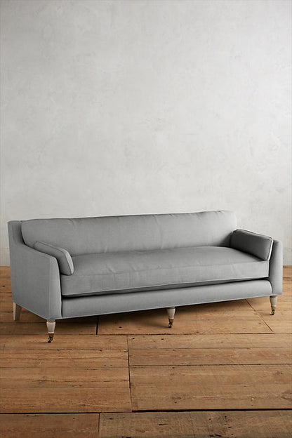 Fotografía de diseño de interiores estilo editorial que muestra un living moderno e industrial sofisticado. En el centro se destaca un sillón de tres cuerpos tapizado en lino gris claro con patas torneadas blancas y rueditas de bronce, idéntico al modelo de referencia. Está ubicado sobre una alfombra de diseño clásico envejecido superpuesta a una gran alfombra de yute
