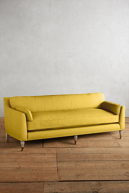 Fotografía de diseño de interiores estilo editorial que muestra un living moderno e industrial sofisticado. En el centro se destaca un sillón de tres cuerpos tapizado en lino gris claro con patas torneadas blancas y rueditas de bronce, idéntico al modelo de referencia. Está ubicado sobre una alfombra de diseño clásico envejecido superpuesta a una gran alfombra de yute
