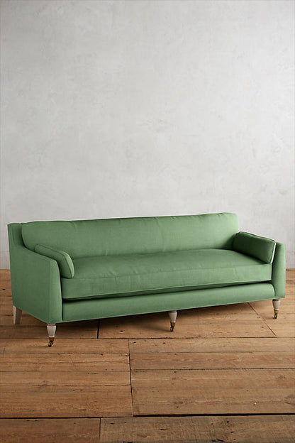 Fotografía de diseño de interiores estilo editorial que muestra un living moderno e industrial sofisticado. En el centro se destaca un sillón de tres cuerpos tapizado en lino gris claro con patas torneadas blancas y rueditas de bronce, idéntico al modelo de referencia. Está ubicado sobre una alfombra de diseño clásico envejecido superpuesta a una gran alfombra de yute