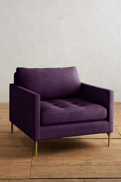 Sillón individual moderno en color violeta con respaldo y asiento acolchados, apoyabrazos rectos y patas metálicas finas en dorado, sobre fondo blanco.