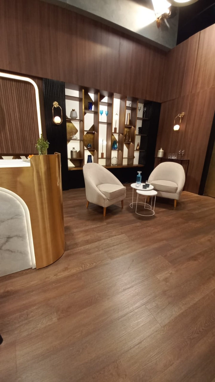 Dos sillones modernos de tapizado beige claro con patas de madera, dispuestos en un área de espera con suelo de madera oscura. Entre ellos hay dos mesas pequeñas redondas, una con una botella y vaso de agua. Al fondo se observa una pared decorativa con estanterías iluminadas y un gran logo dorado de MasterChef