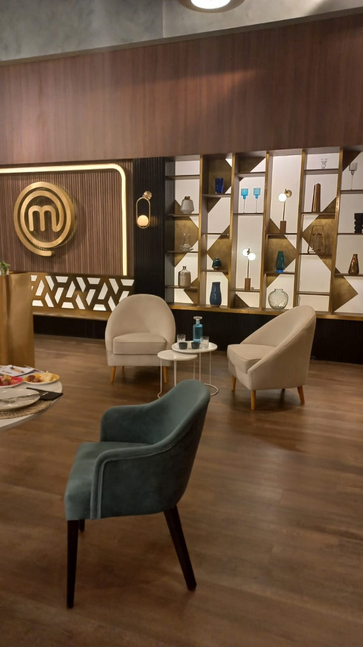 Dos sillones modernos de tapizado beige claro con patas de madera, dispuestos en un área de espera con suelo de madera oscura. Entre ellos hay dos mesas pequeñas redondas, una con una botella y vaso de agua. Al fondo se observa una pared decorativa con estanterías iluminadas y un gran logo dorado de MasterChef