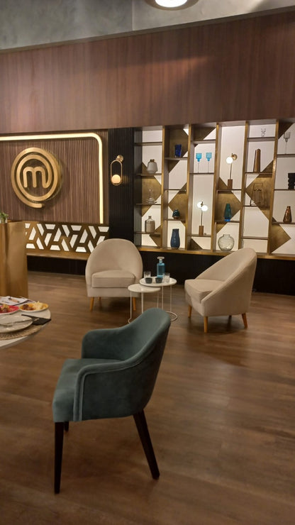Dos sillones modernos de tapizado beige claro con patas de madera, dispuestos en un área de espera con suelo de madera oscura. Entre ellos hay dos mesas pequeñas redondas, una con una botella y vaso de agua. Al fondo se observa una pared decorativa con estanterías iluminadas y un gran logo dorado de MasterChef