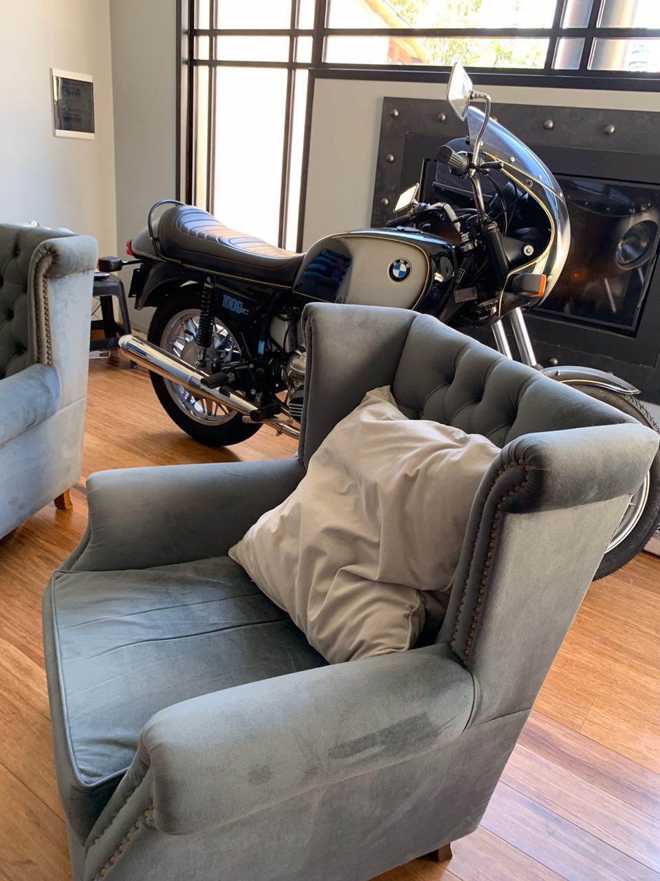 Sillón de un cuerpo color gris topo con capitoné y una moto estilo vintage de fondo 